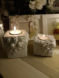 two white candles sitting on top of a table next to some flowers and framed pictures