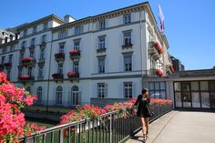 a woman walking down a sidewalk next to a tall white building with red flowers in front of it