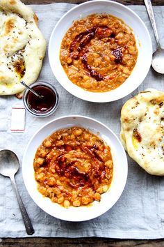 three bowls filled with food on top of a white table cloth next to bread and spoons