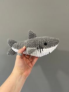 a hand holding a gray and white knitted shark toy in front of a grey wall