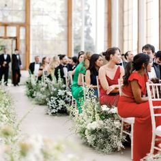 two women in red dresses sitting next to each other on white chairs with flowers and greenery