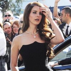 a woman in a black dress is standing next to a car and people are looking at her