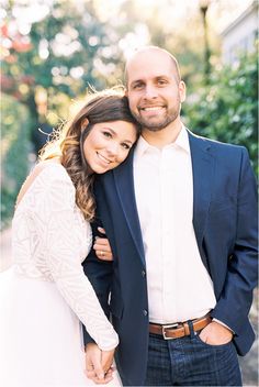 a man and woman standing next to each other in front of some green trees with their arms around each other