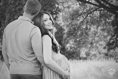 a pregnant woman standing next to a man in front of a tree and grass field