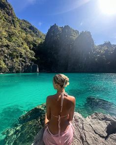 a woman sitting on the edge of a cliff looking out at blue water and mountains