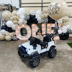 a little boy is sitting in a toy car with balloons and decorations behind him that spell out one happy dude