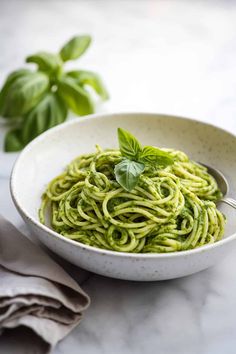 a white bowl filled with pesto pasta on top of a marble table next to basil leaves