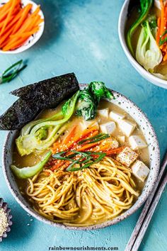 two bowls filled with noodles, vegetables and tofu on top of a blue table