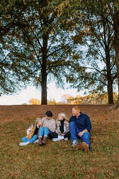 three people sitting on the ground in front of trees with leaves all around them and one person holding a dog