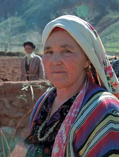 an old woman with a scarf on her head standing in front of other people and looking at the camera