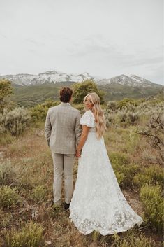 a bride and groom holding hands walking through the grass with mountains in the background on their wedding day