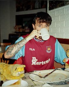 a young man sitting at a table drinking from a cup with stuffed animals around him