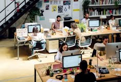 people working on their computers in an office with bookshelves and stairs leading up to the second floor