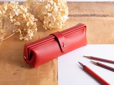 a red leather pencil case sitting on top of a wooden table next to some flowers