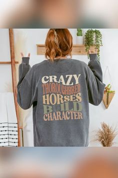 A woman facing away from the camera, wearing a dark gray sweatshirt with bold, colorful text reading, 'CRAZY THOROUGHBRED HORSES BUILD CHARACTER.' She holds up peace signs with both hands, and her hair is tied in a loose ponytail. The background includes wall-mounted plants, a wooden shelf, and a cozy, bohemian decor setting. Equestrian Tshirts, Horse Trainer Gifts, Cowgirl Tshirt, Horse Trainer, Thoroughbred Horse, Horse Shirt