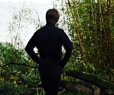 a man standing in front of bamboo trees and looking at the water with his hands on his hips