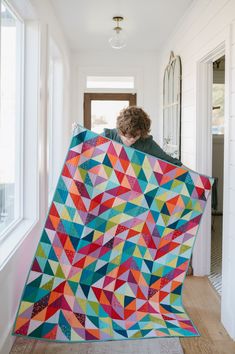 a woman holding up a colorful quilt in front of a window with white walls and wood flooring