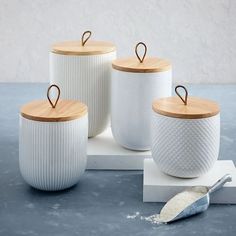 three white canisters with wooden lids are on a table next to a spoon