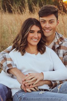 a man and woman sitting on the ground in front of tall grass with their arms around each other