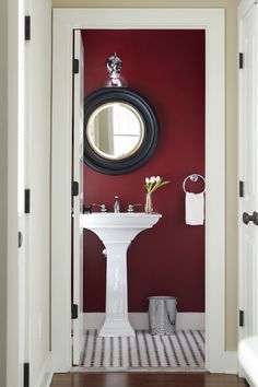 a bathroom with red walls and a white pedestal sink under a round mirror on the wall