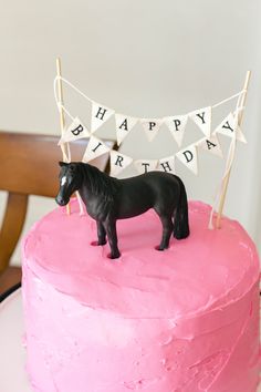 a pink birthday cake with a horse figurine on top and happy birthday banner