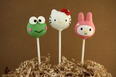 three cake pops with hello kitty, frog and bunny faces on top of shredded paper