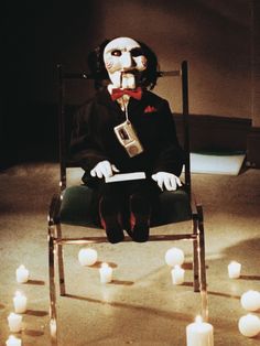 a creepy doll is sitting in a chair with candles around him and his face painted white