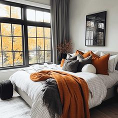 a bed with orange and gray pillows in front of a large window, next to a wooden floor