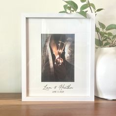 a white vase sitting on top of a wooden table next to a framed photo and a plant