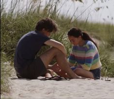 a man and woman sitting on top of a sandy beach next to tall green grass