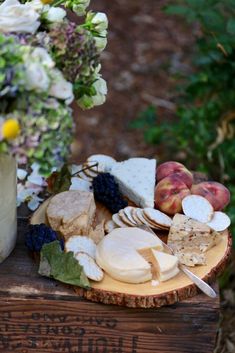 an assortment of cheeses and fruit on a wooden table outside with flowers in the background