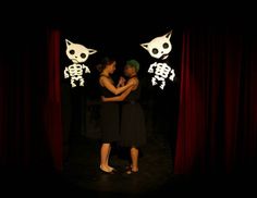 two women standing in front of red curtains with skeleton decorations on the wall behind them