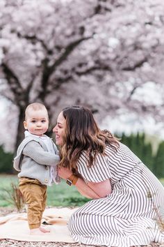 a woman holding a baby in her arms while sitting on a blanket with cherry blossom trees in the background