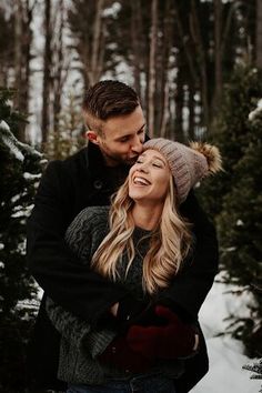 a man and woman standing in the snow hugging each other with trees in the background