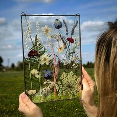 a woman holding up a glass plate with flowers and plants on it in front of a field