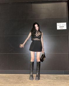 a woman standing in front of a wall holding a handbag and wearing black boots