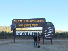 two people standing in front of a sign for napa valley wine country, with mountains in the background