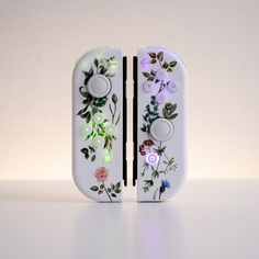 two video game controllers sitting side by side on top of a white surface with flowers and leaves painted on them