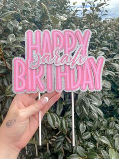 a hand holding two pink happy birthday cake toppers in front of some bushes and trees