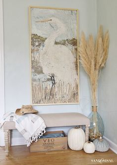 a painting hangs on the wall next to a bench and vase with dried grass in it