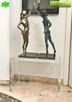 two bronze statues are sitting on a glass table in front of a framed art piece