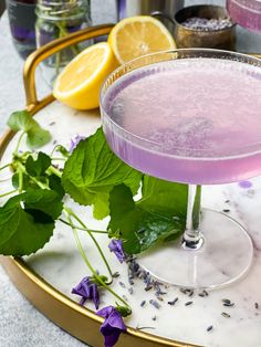 a purple drink in a glass on a tray next to lemons and lavender sprigs