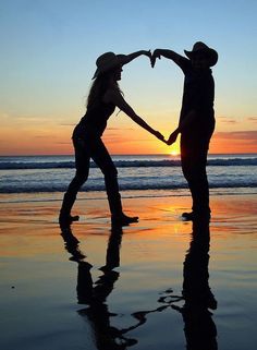 two people standing on the beach holding hands and making a heart shape with their hands