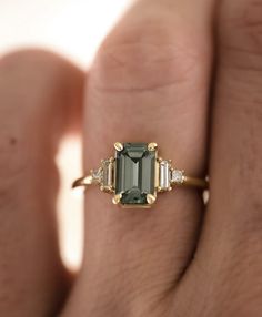 a woman's hand holding a ring with an emerald stone and three diamond baguets