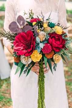 a bride holding a bouquet of red and yellow flowers with greenery in her hands