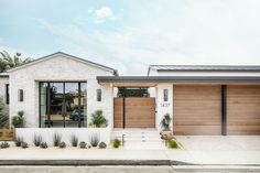 a white brick house with wood garage doors and windows on the front side, along with plants and trees