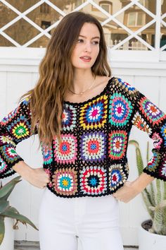 a woman standing in front of a house wearing white pants and a colorful crochet sweater