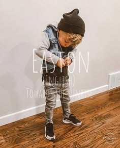 a young boy standing on top of a hard wood floor wearing a black and gray hat