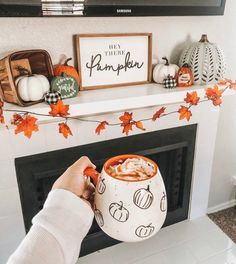 a person holding a mug in front of a fireplace with fall decorations on the mantle