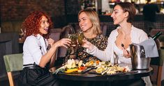 three beautiful women sitting at a table with wine glasses in their hands and food on the plate